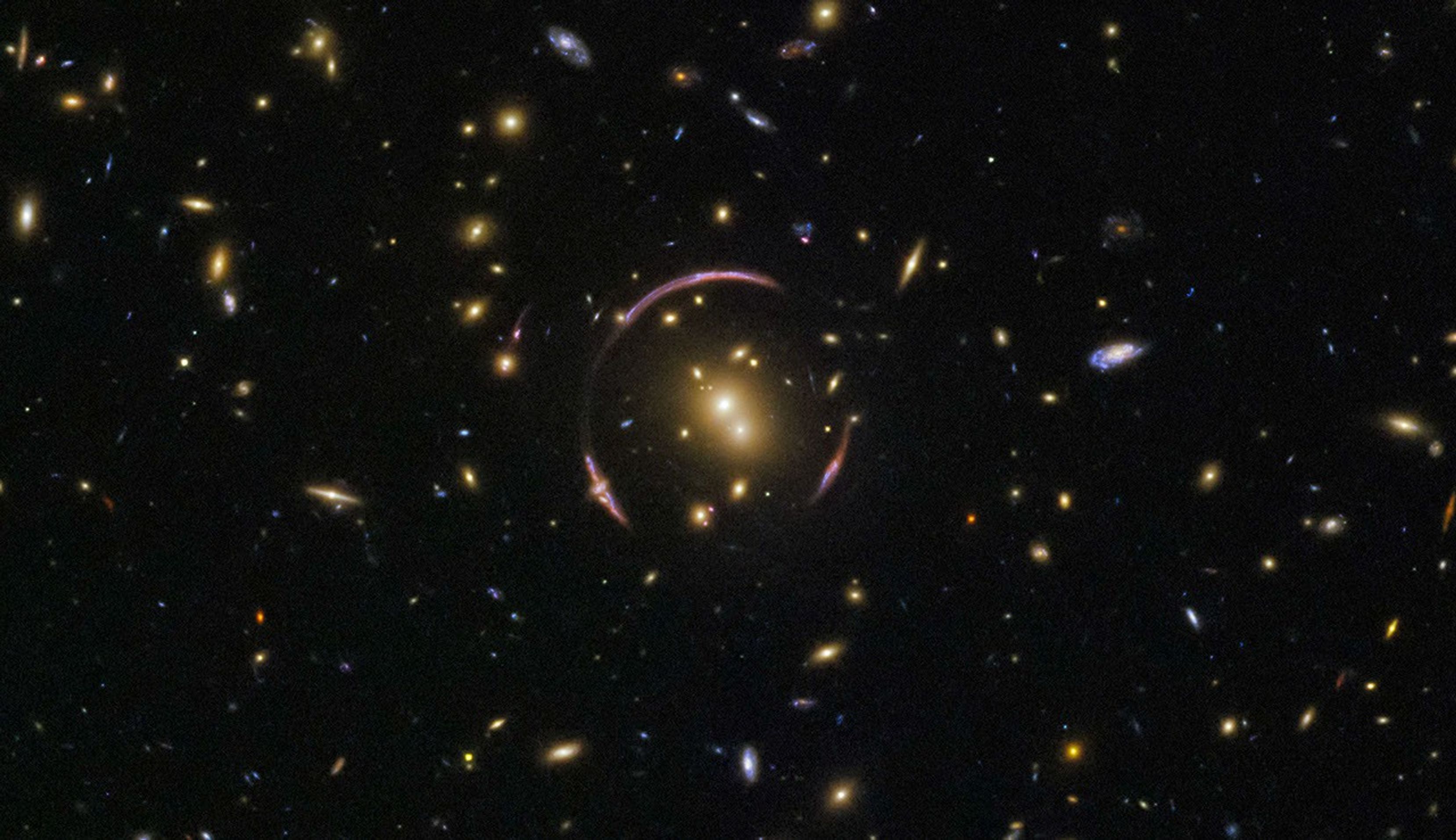 Black background dotted with galaxies. A reddish-orange pair of galaxies at image center with a pinkish-red ring curving almost completely around it.